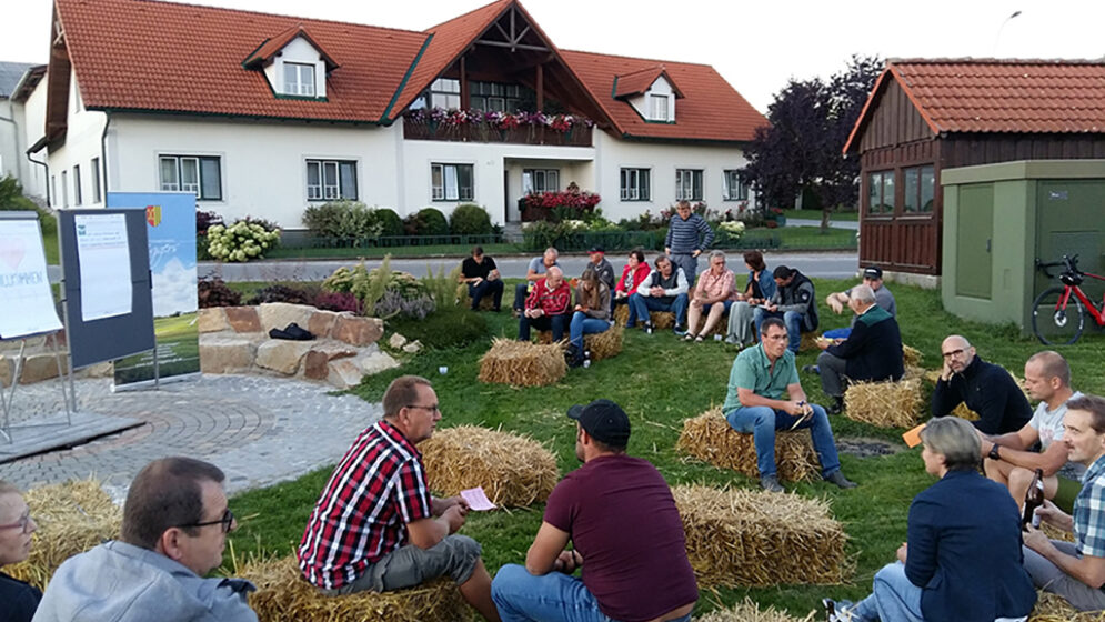 Am Fotos sind auf verstreut liegenden Strohballen sitzende Menschen zu sehen, die einem Vortragenden zuhören.